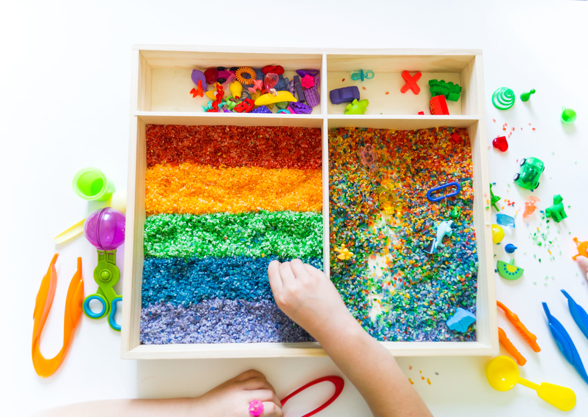 A child&rsquo;s hand exploring a rainbow sensory rice tray